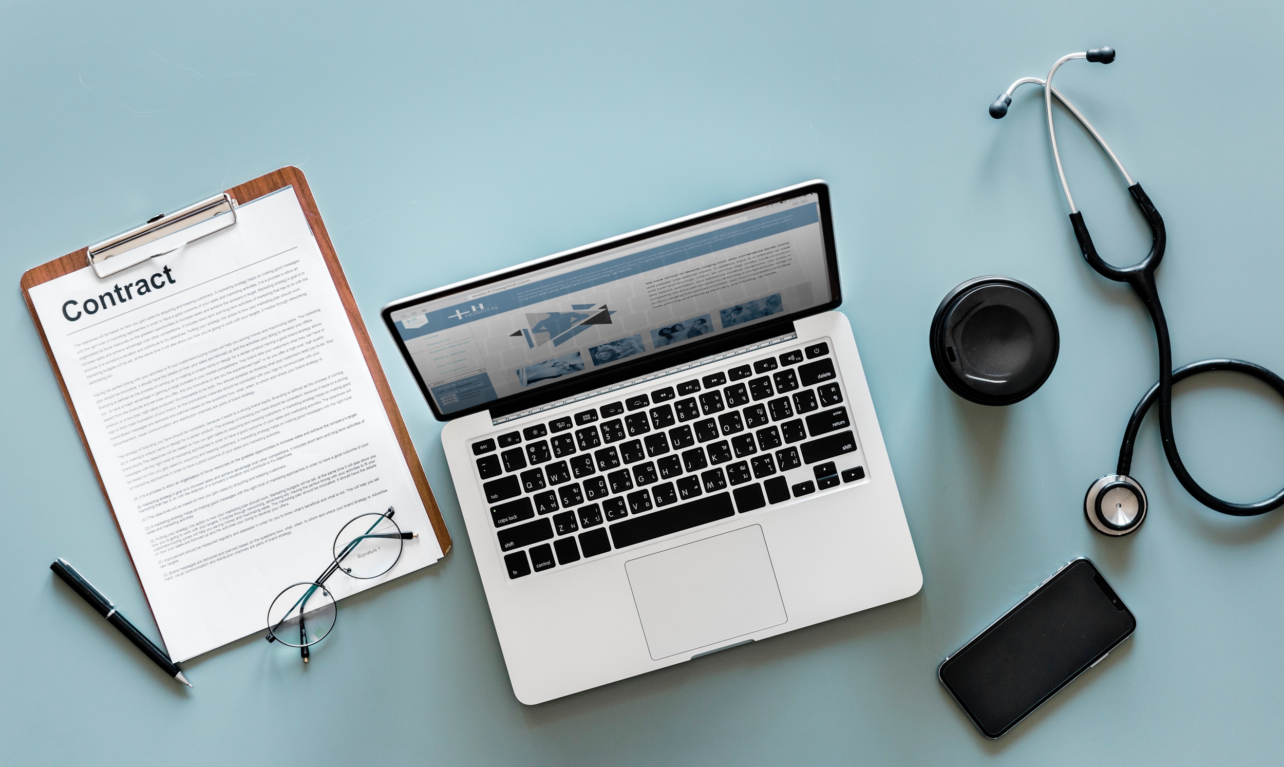 photograph of a pen, contract, reading glasses, laptop, phone, and stethoscope